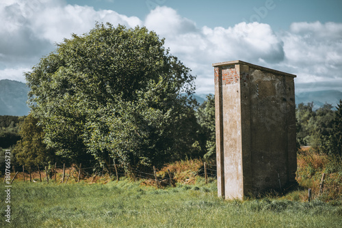 Фотография Weathered concrete tower standing in a lush green meadow beside dense trees under bright clouds and distant mountains, creating a rustic rural scene with strong textures and natural light