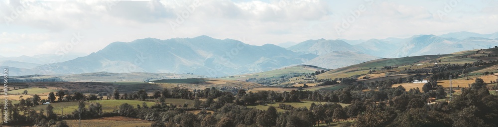 Fototapeta premium Wide telephoto panorama of rolling green hills, patchwork farmland and distant rugged mountains in soft light along Camino de Santiago in Asturias, capturing serene rural landscape beauty