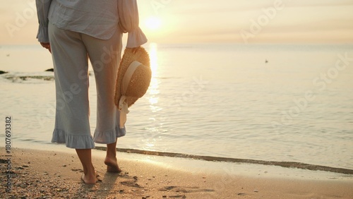 Walking along a serene beach at sunset while holding a straw hat, enjoying the tranquility of nature and the gentle waves