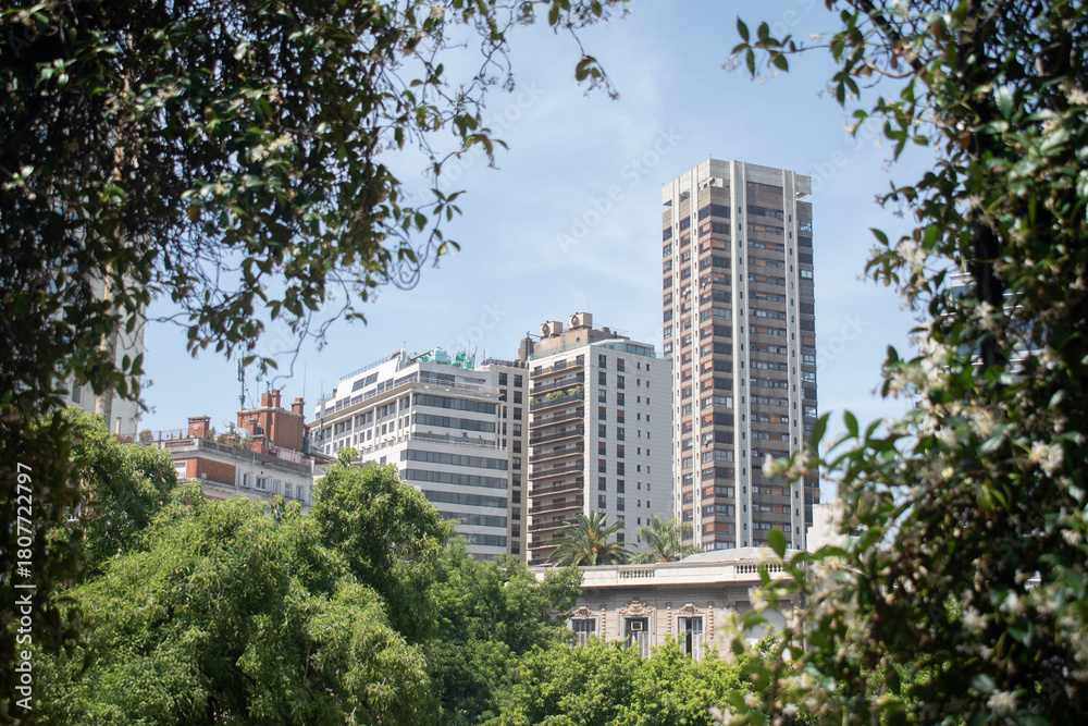 Fototapeta premium Panoramic view of modern high-rise residential and office buildings framed by lush urban greenery on a bright sunny day.
