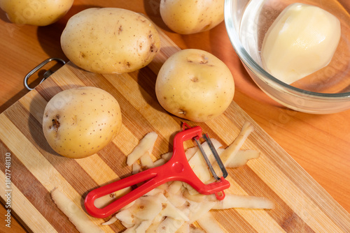 Unpeeled and peeled potatoes and a red vegetable peeler on a kitchen cutting board. Top-down view showcasing the process of food preparation: peelings cut with a peeler and a peeled potato
