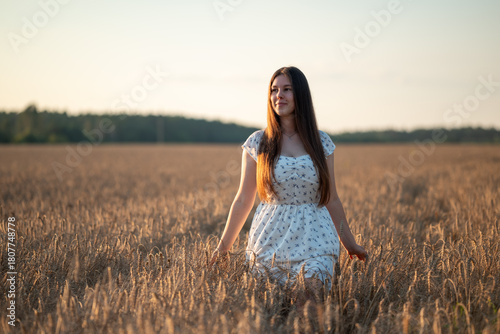 Girl in a light dress standing in a wheat field at sunset. A young woman in a white dress standing among golden wheat ears, looking into the distance, harmony, calmness, connection to nature