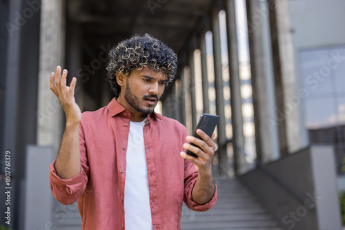 A worried young Indian man stands on the street outside an office building and stares in dismay at the screen of the phone he holds in his hand