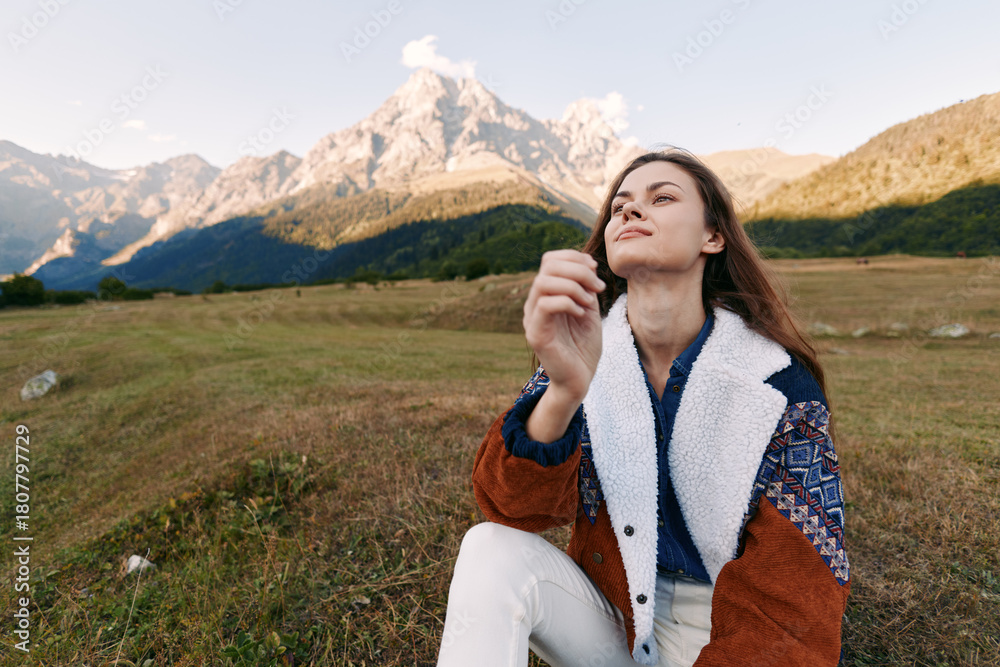 Fototapeta premium Woman portrait in meadow by mountains, nature outdoors scene showing a hiking break and relaxation, female traveler in warm jacket enjoying alpine landscape and clear sky.
