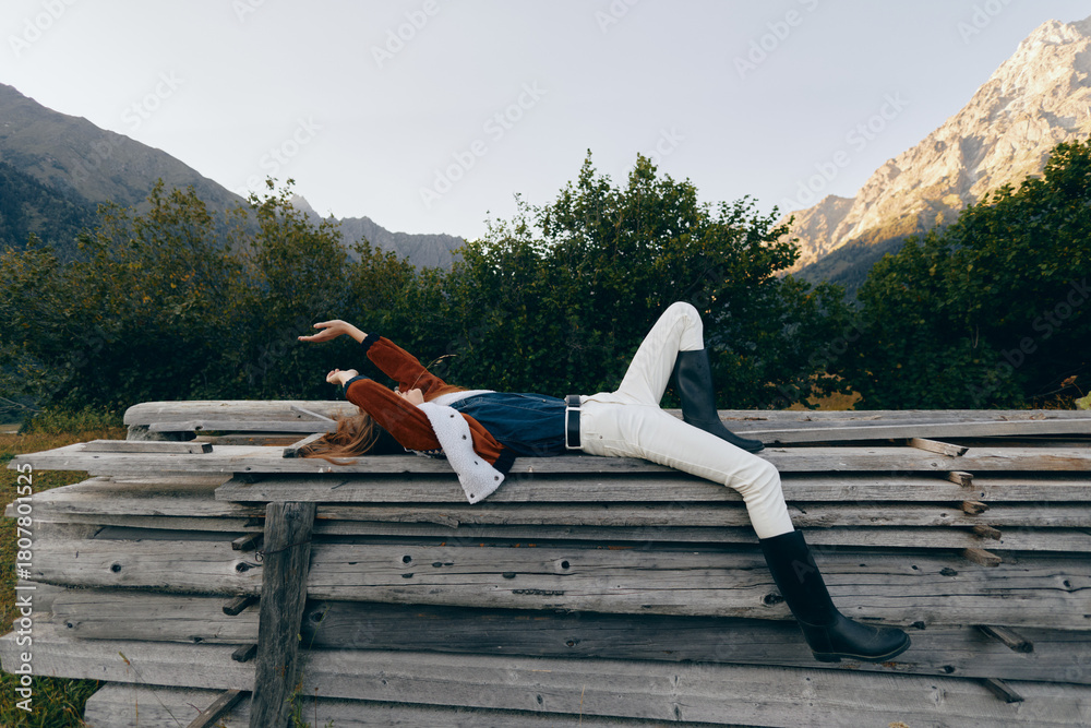 Fototapeta premium Woman lying on stacked logs in mountain landscape, outdoors relaxation and leisure with denim jacket, white pants and black boots, stretching arms back under clear sky.