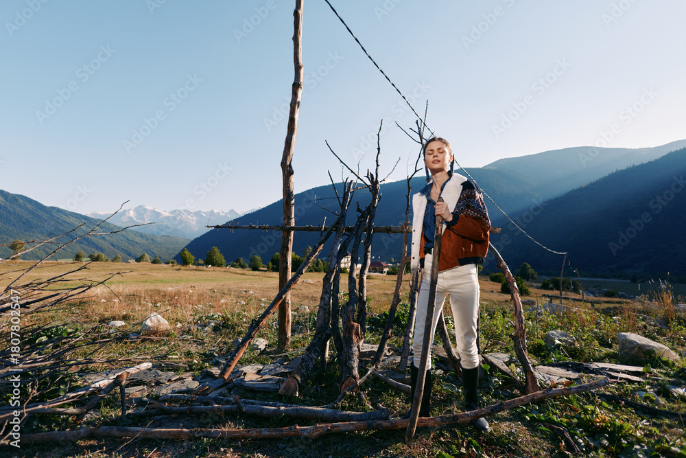 Fototapeta premium Woman portrait and traveler standing by rustic wooden fence in meadow below mountains, landscape and nature scene with clear sky, sunlight and autumn fashion jacket and boots.