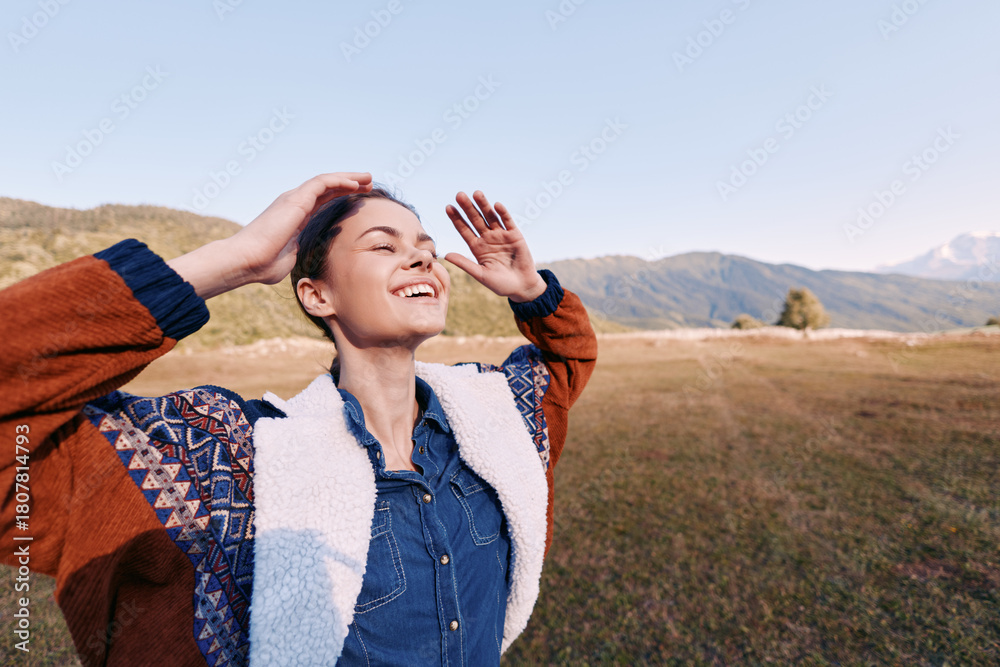 Fototapeta premium Woman smiling with eyes closed outdoors in countryside, hands raised enjoying nature and warm sunshine, happiness and freedom in casual jacket and denim, relaxed joyful portrait in meadow.