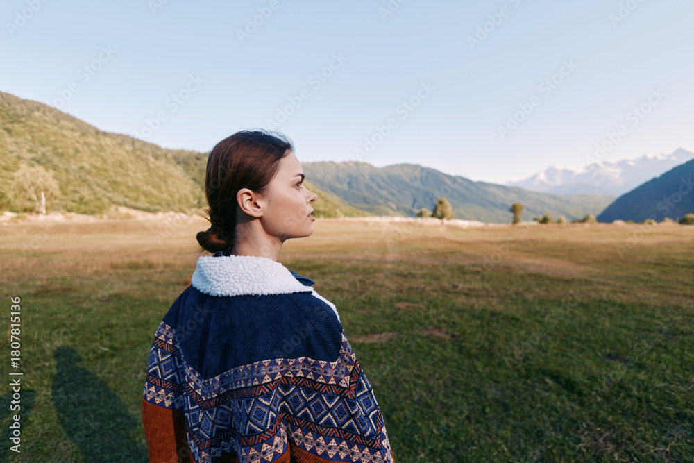 Fototapeta premium Woman in patterned sweater stands in meadow looking at distant mountains, profile view in outdoor landscape. Nature travel scene with calm light, open field and valley retreat.