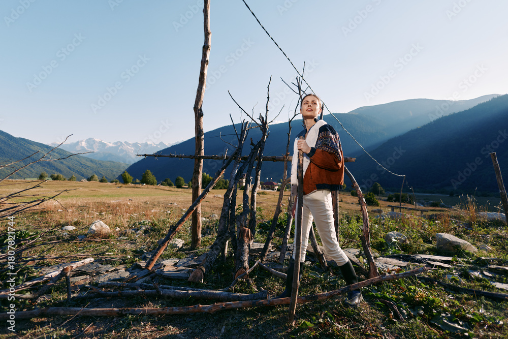 Fototapeta premium Woman hiking with backpack in mountain meadow, standing near rustic wooden fence, enjoying nature and outdoors travel, scenic landscape with distant peaks and clear blue sky.