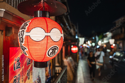 Tableau sur toile A red and white chochin with bold kanji hangs by a wooden shop in Kyoto, with machiya facades and lanterns along a narrow lane as people stroll in soft focus
