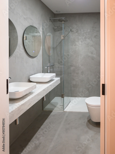 A contemporary bathroom features microcement walls and floors, double sinks, and a glass shower, embracing a modern minimalist aesthetic that highlights durability and seamless design.