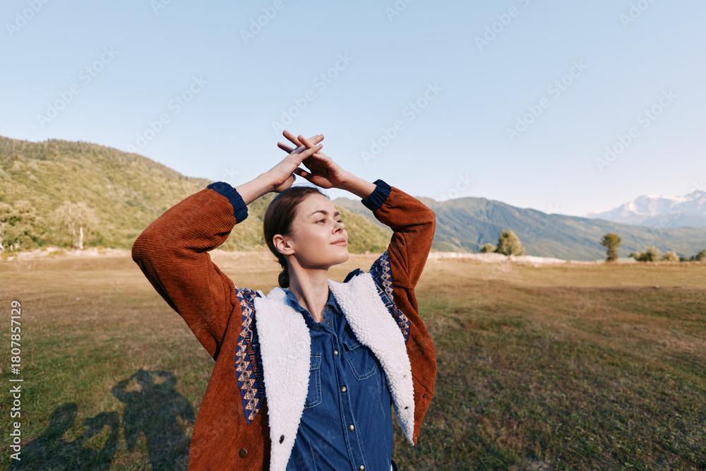 Fototapeta premium Woman portrait in nature among mountains and open field, relaxed and eyes closed outdoors wearing a cozy sweater, enjoying sunlight, breeze and peaceful rural scenery on a sunny day.