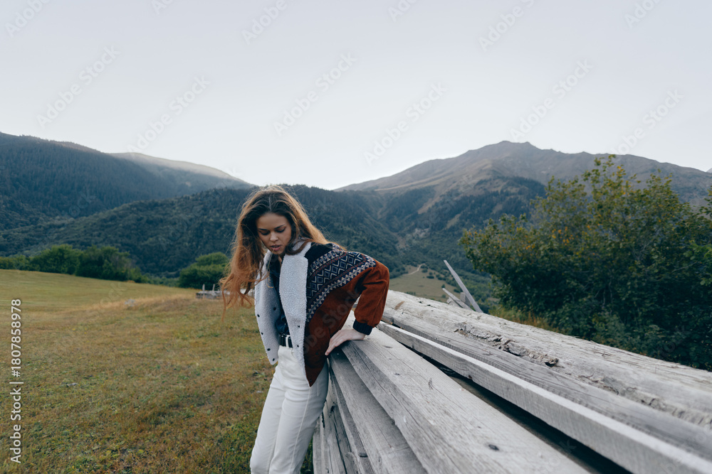 Fototapeta premium Woman leaning on wooden fence in meadow with mountains background, wearing patterned jacket and white pants. Outdoor portrait, countryside landscape and moody natural scenery.