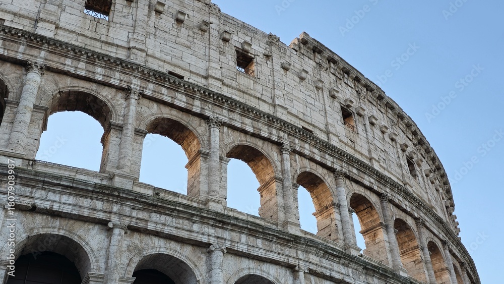 Fototapeta premium Rome, Italy - 13 January 2025. The Colosseum’s upper arches are lit by warm sunlight under a clear blue sky, captured from a low angle.