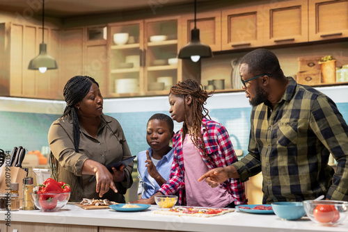 Smiling children following online recipe for pizza with their parents, preparing homemade food step by step with fresh ingredients. Black family gathered around the kitchen counter.
