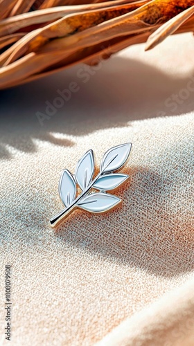 A close-up shot of a small, white leaf branch pin with gold accents, placed on a textured fabric surface, with dried plant material and soft light in the backgr