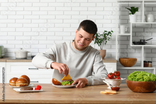Smiling young man preparing homemade burgers in modern kitchen, adding cheese...