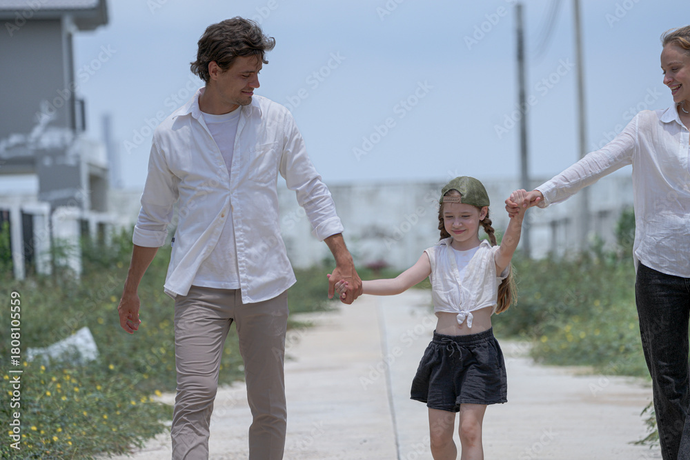 Fototapeta premium A cheerful family walks together through their new neighborhood. The smiling parents hold their daughter's hands, lifting her playfully as they enjoy a happy walk in front of their home.