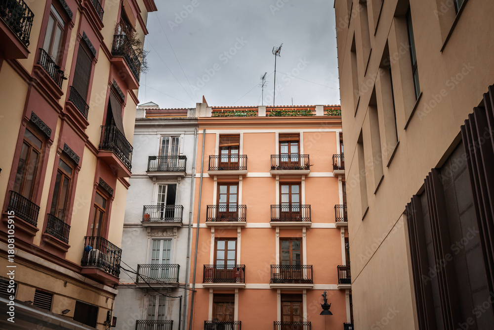 Obraz premium Residential apartment buildings with pastel facades and balconies face a narrow street in Ciutat Vella, the historic old town of Valencia, Spain, illustrating urban housing and local real estate.
