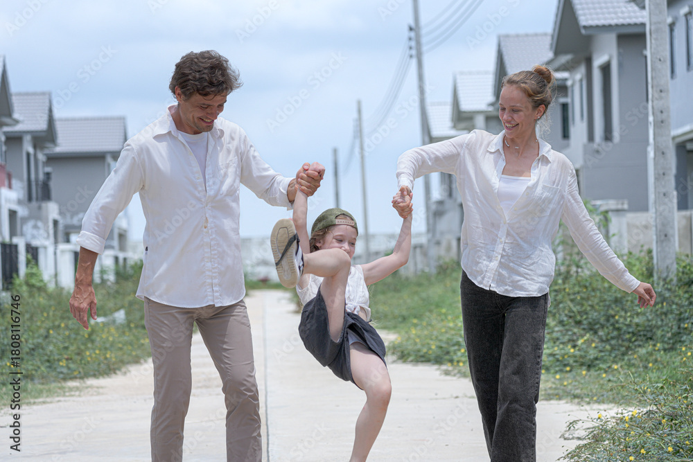 Obraz premium A cheerful family walks together through their new neighborhood. The smiling parents hold their daughter's hands, lifting her playfully as they enjoy a happy walk in front of their home.
