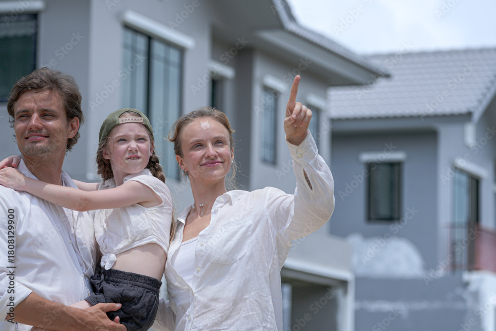 Obraz premium A hopeful young family looks towards their future outside a new modern home. The father holds their daughter, who points excitedly, as the mother smiles, dreaming of their life together.