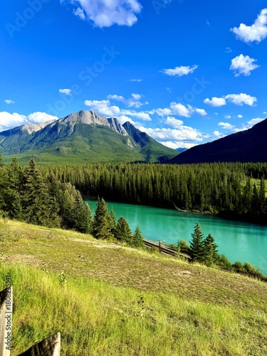 Scenic view of a tranquil turquoise river winding through lush green forested mountains under a bright blue sky with fluffy white clouds, showcasing natural beauty and serenity