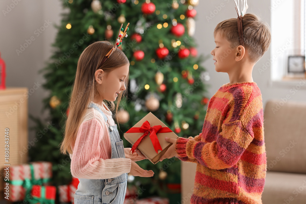 Naklejka premium Cute little boy giving his sister book at home on Christmas eve