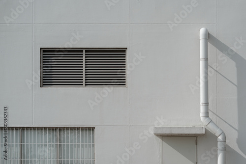 Industrial white wall facade featuring a louvered vent, barred window, and a vertical drainage pipe.