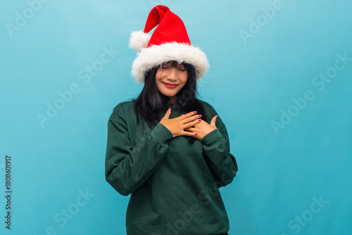 Asian woman in Santa hat touches her chest smiling warmly Ideal for heartfelt Christmas greetings and cozy visuals