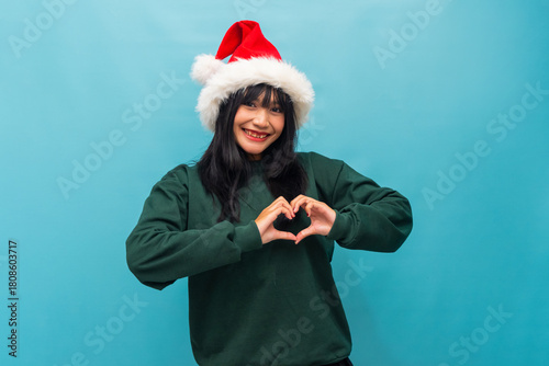Happy Asian woman in Santa hat forms a heart shape with hands Ideal for Christmas love messages and holiday ads