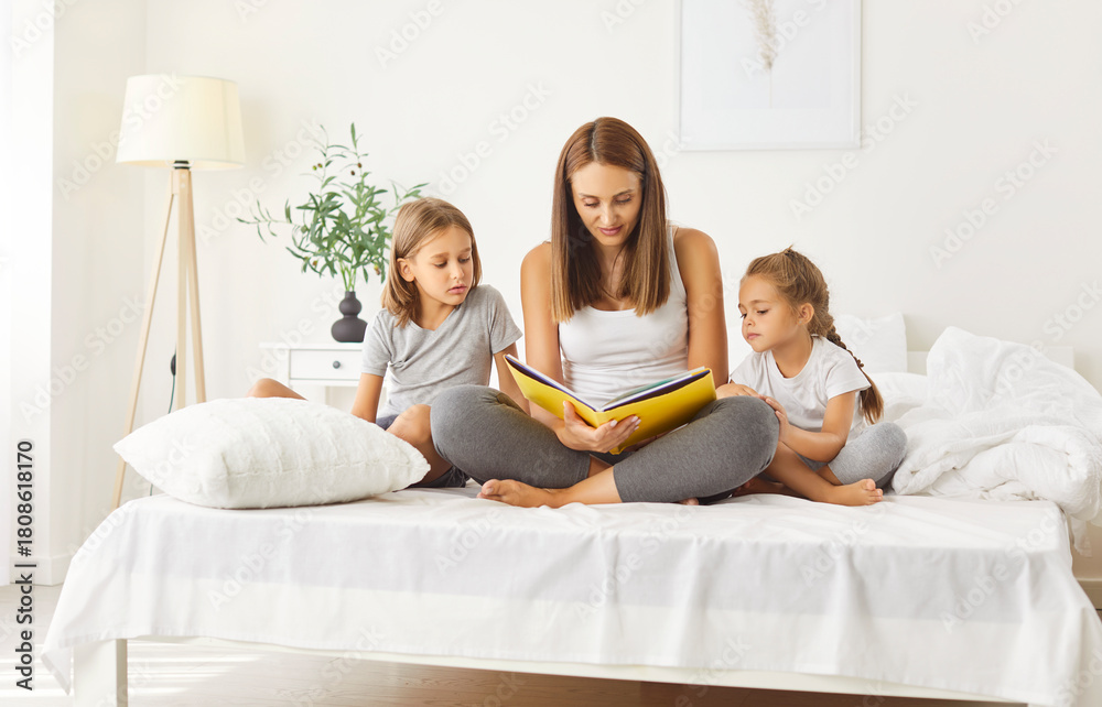 Fototapeta premium Young mother sitting on bed with her little children and reading them fairytale. Caring mom holding book, enjoying storytelling with two kids at home. Happy family spending leisure time together.