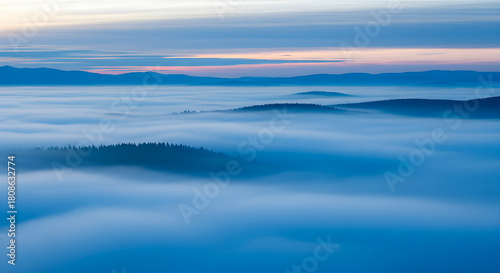 Breathtaking aerial view of rolling hills and forests submerged under a thick blanket of blue fog or low clouds, with mountain ridges peaking through.
