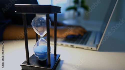 Close-up of an hourglass with blue sand on a desk, person typing on a laptop in the background