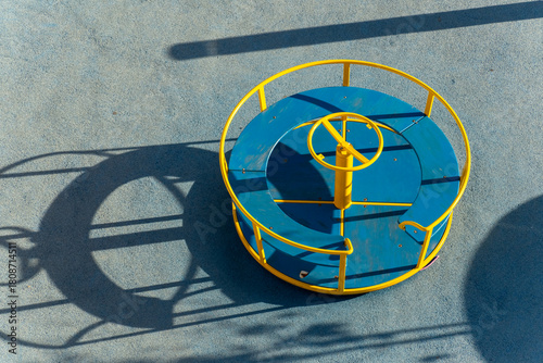 Close-up view of a blue and yellow playground roundabout casting bold circular shadows onto a textured blue surface