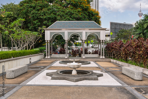 Modern Islamic Pavilion and Fountain at National Mosque, Kuala Lumpur, Malaysia. KL Tower in background, surrounded by lush tropical gardens under a cloudy sky