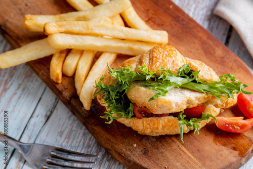 chicken croissant sandwich served with a side of french fries on a wooden board. The sandwich is made with a flaky croissant and filled with a piece of chicken, green lettuce, and sliced red tomatoes.