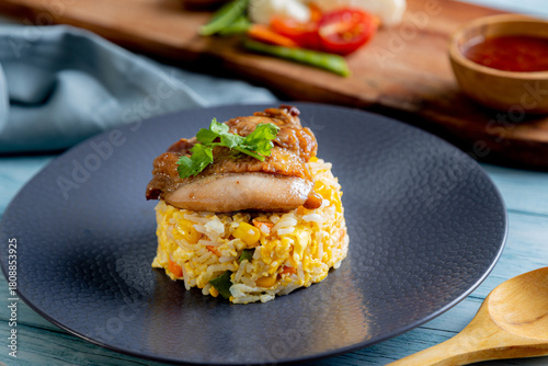 A serving of fried rice with corn and egg is topped with a piece of grilled chicken and a sprig of cilantro, presented on a dark plate. wooden board holds a small bowl of holds a a  sauce, vegetables
