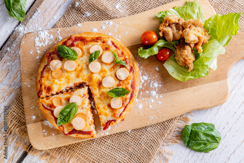 freshly baked sausage pizza with basil on a wooden cutting board, with one slice removed. Alongside the pizza on the same board is a side serving of crispy fried chicken pieces resting on lettuce