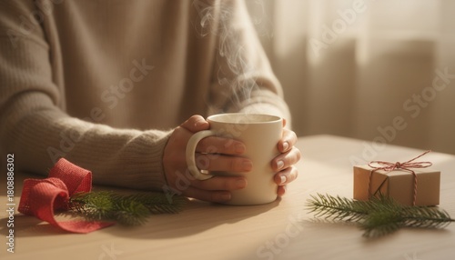Woman hands cupping a hot beverage mug on a table with red ribbon and gift. Cozy winter season and holiday celebration concept.