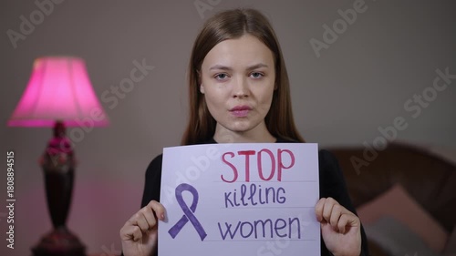 White woman holding stop killing women sign in dim living room with pink lamp, steady gaze as activist, survivor advocate and community organizer making direct
