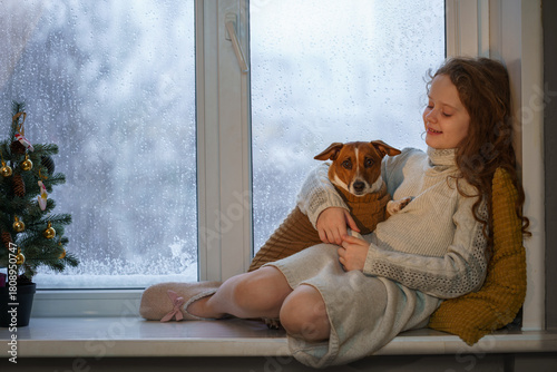 A young girl and a Jack Russell Terrier dog share a cozy moment on a windowsill, watching the snow fall outside. Christmas time.