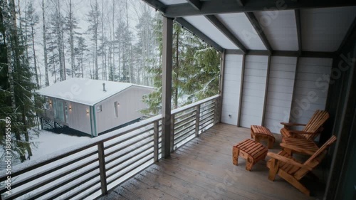 No people shot of wooden porch with rustic chairs overlooking snow covered forest and small cabin creating peaceful winter retreat atmosphere