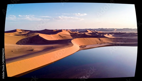 Fototapeta Naklejka Na Ścianę i Meble -  A tranquil desert landscape featuring vast sand dunes bordering a still body of water that mirrors the sandy formations and the sky above.