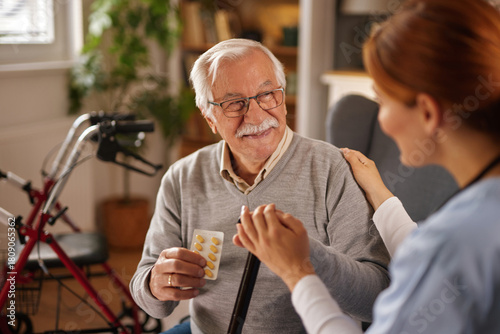 An elderly man with grey hair and glasses smiles while holding a blister pack of pills and a cane. A nurse places her hand on his shoulder, offering support and reassurance in his home.