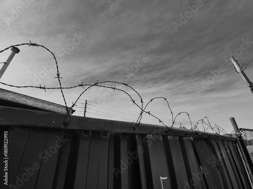 Black and white close-up of barbed wire coiled around the top edge of a fence, against a whispy cloudy sky.