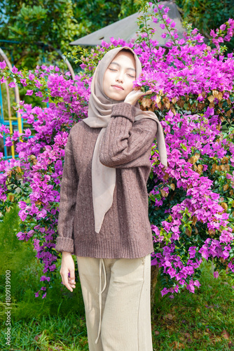 A Muslim Asian woman appears graceful and peaceful as she poses for a photo, kissing a lush purple bougainvillea flower. The beautiful woman poses against a backdrop of pink paper flowers.