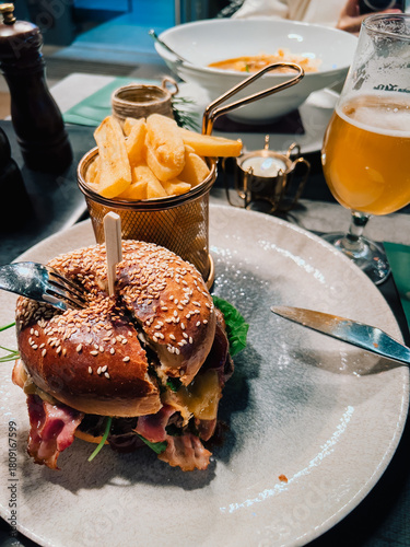 Juicy burger with bacon, cheese, and fresh arugula is being enjoyed with side of crispy fries and refreshing glass of beer, creating satisfying dining experience. Juicy bacon cheeseburger, fries, beer