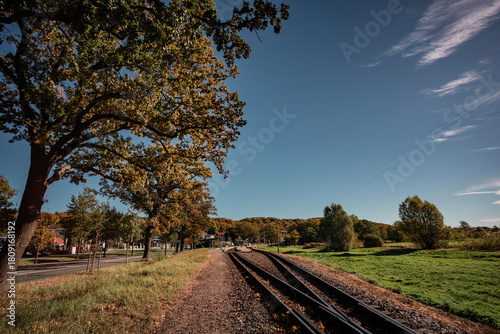 Perspective view of the narrow-gauge railway tracks leading into the distance, lined by trees with early autumn foliage under a dramatic blue sky with wispy clouds.