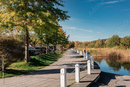 Paved autumn pathway along a pond or small lake, lined with trees and a bollard chain fence. Parked cars are visible on the left, with two people enjoying the view.