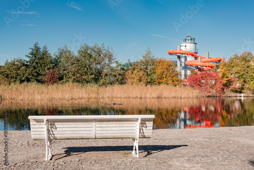 A white bench overlooks a pond reflecting autumn trees and the tower and orange water slides of a modern spa or leisure facility in the background.